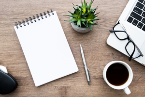 image of a blank notepad sitting on a wooden desk next, accompanied by a small plant, a pen, a cup of coffee, and a laptop computer with glasses lying on it