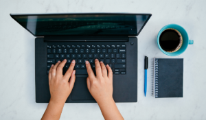 two hands on a laptop keyboard, next to a pen, a notebook, and a cup of coffee