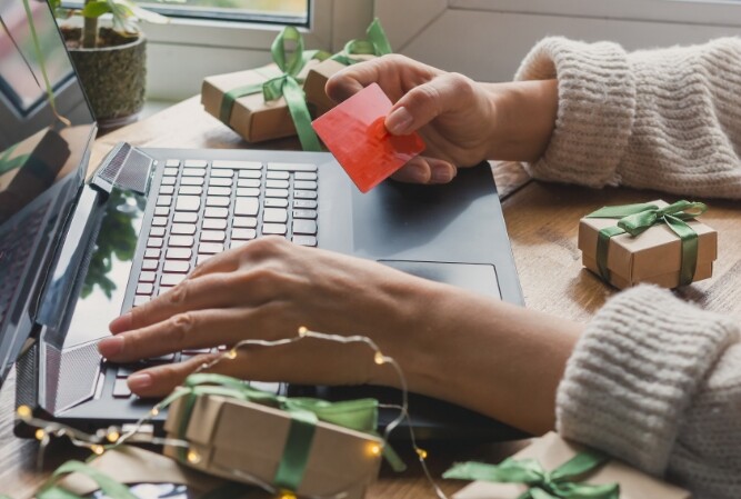 a woman in a sweater holds a credit card while sitting at her computer, decorated with red and green